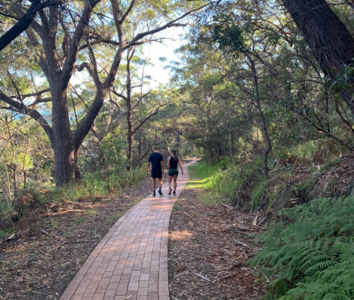 A couple in activewear walk down a red brick path through a coastal forest, with blue sea glimpsing between the trees