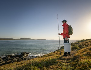 Fisherperson standing with fishing rod and bucket looking towards the sea