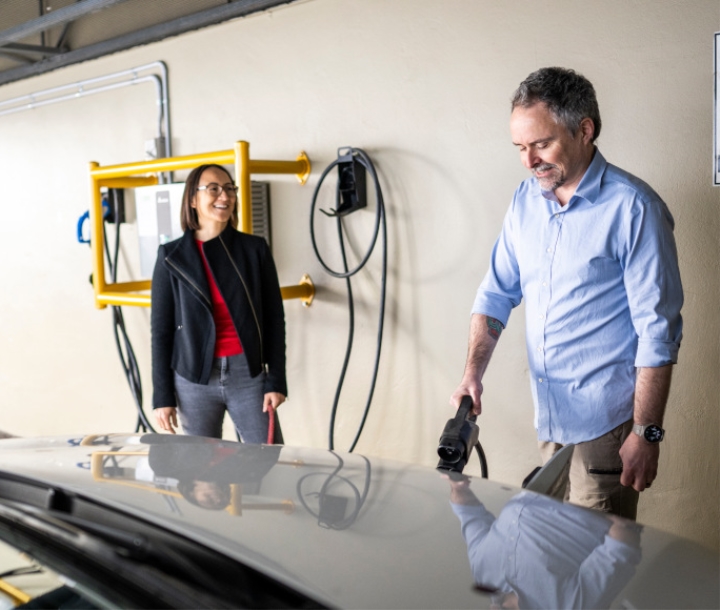 Smiling man charging an electric vehicle in an apartment building that has been retrofitted with a charging station, next to him is a woman with a whippet or greyhound on lead but just out of shot