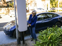 Woman in blue with Eastern suburbs hairstyle posing by blue car and EV charger with her phone out