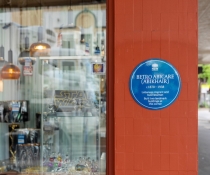 Blue plaque for Betro Abicare on a terracotta coloured shopfront. The shop window is filled with vintage glass pieces and the shop is on a corner under an awning.