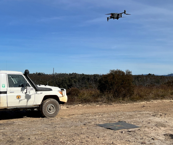 A dirt road with a National Parks and Wildlife Service truck parked on it, and a black drone overhead; dark brown-green scrub in the background