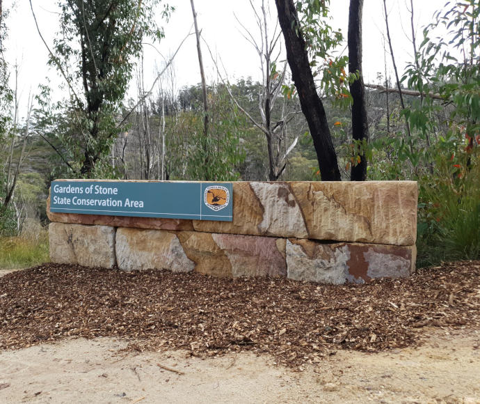 A sign announcing the 'Gardens of Stone State Conservation Area' on a low stone wall with the National Parks and Wildlife Service logo of a black lyrebird over an orange background, with tanbark in front of the sign and trees  behind