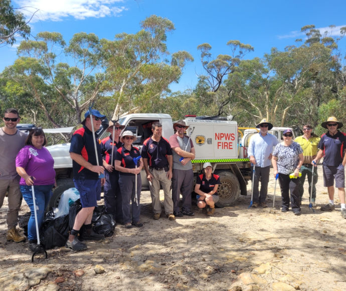 An assemblage of people of different ages, wearing hats and sunglasses, and each holding a litter-grabbing stick, posed in front of a National Parks and Wildlife Service truck with a bright blue sky and verdant eucalypt foliage in the background