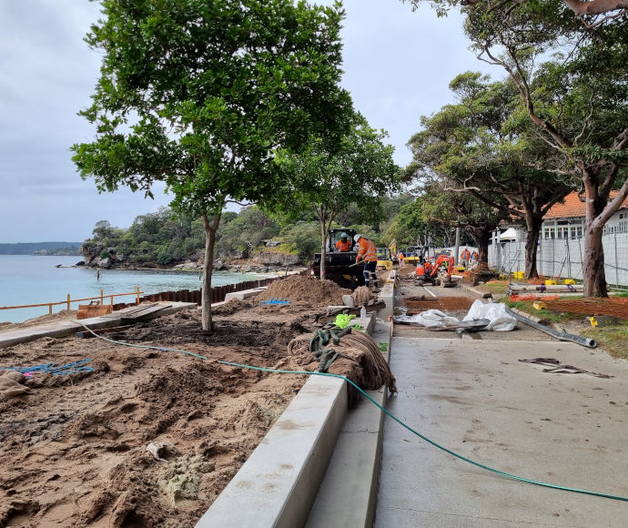 Workmen and saplings in fresh dirt along a promenade under construction, overlooking a small picturesque bay