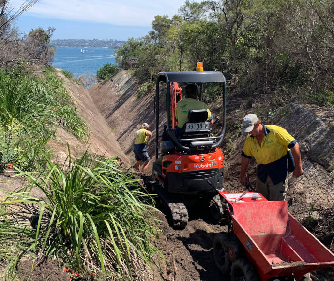 Workmen with red excavation equipment remove dirt from an old military ditch, with greenery lining its upper edges and the blue water of Sydney Harbour behind them