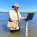 Man wearing a hat and satchel bag, up to his thighs in water, logging data on a laptop resting on a depth logger, Macquarie Marshes