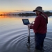 Man in knee-deep water working on laptop on depth logger, sun on the horizon, Macquarie Marshes