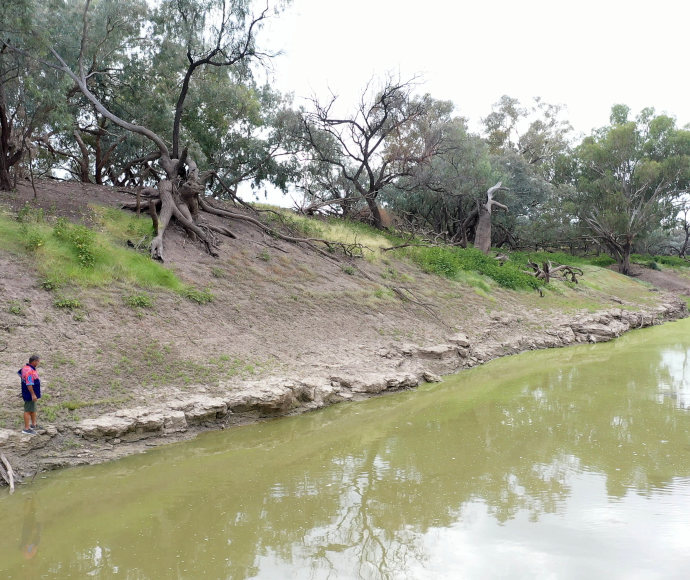 Fred Hooper, standing at the edge of the still, brown river, surveying it, a steep river bank with trees behind him