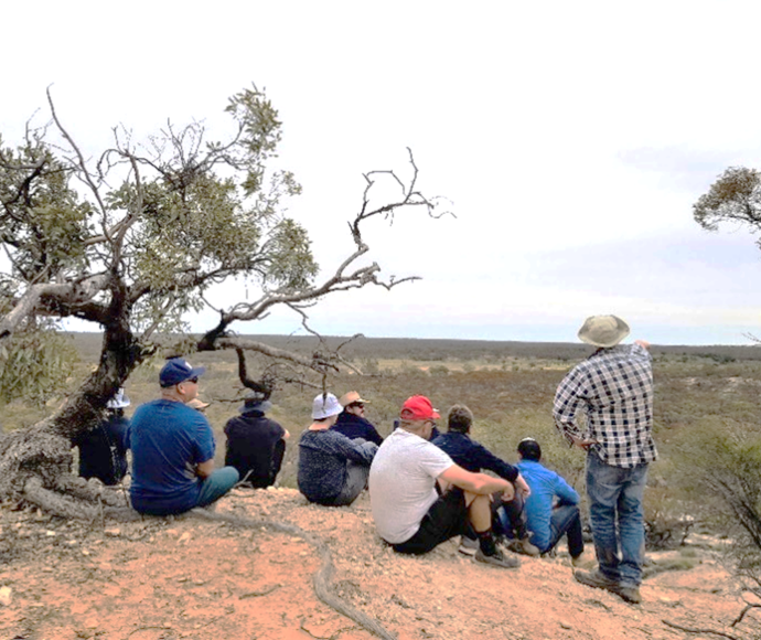 A group of casually dressed staff in hats, sitting next to a scrubby tree on a sandy hill. A man in a floppy hat and checked shirt is standing and pointing to the landscape beyond.