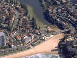 Aerial view of Manly Lagoon, showing a beachfront with waves crashing onto the shore, a river meandering through the landscape, residential areas with numerous houses, greenery, and roads, and a clear demarcation where the urban area meets the natural water bodies.