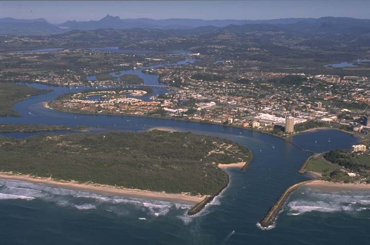 Aerial view of Tweed River flowing into the sea with a breakwater on one side, surrounded by urban development amidst green landscapes with hills in the distance under a clear sky.