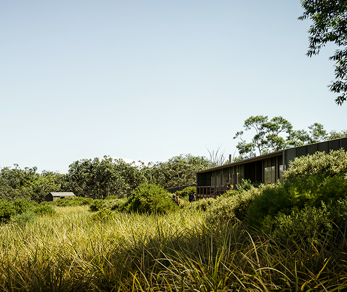 "Modern house with metal roof and smoking chimney, surrounded by lush green trees. In the background, a body of water and distant structures are partially visible through the foliage.