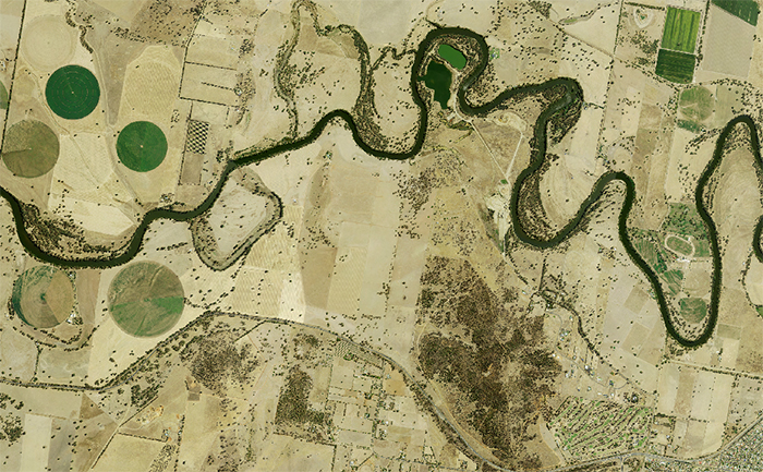 Aerial view of a diverse landscape showing a winding river, various vegetation patterns including circular irrigated fields, dense tree clusters, and scattered shrubbery. The image also depicts different land uses such as agricultural plots and built-up areas, illustrating the complexity of land cover in a given region