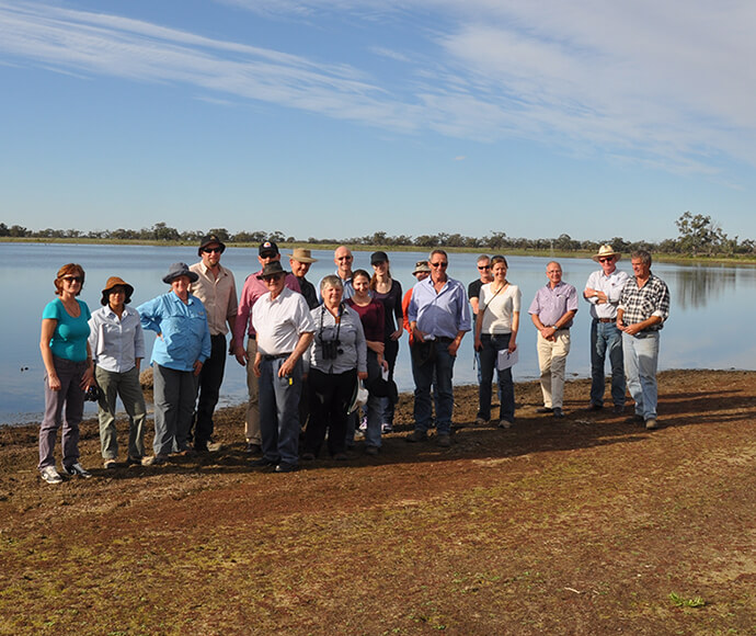 Group photo of the Murrumbidgee EWAG committee at Hobbler's Lake. The group is standing on a dirt ground with sparse greenery, facing the camera. In the background, there is a calm lake and a clear sky.