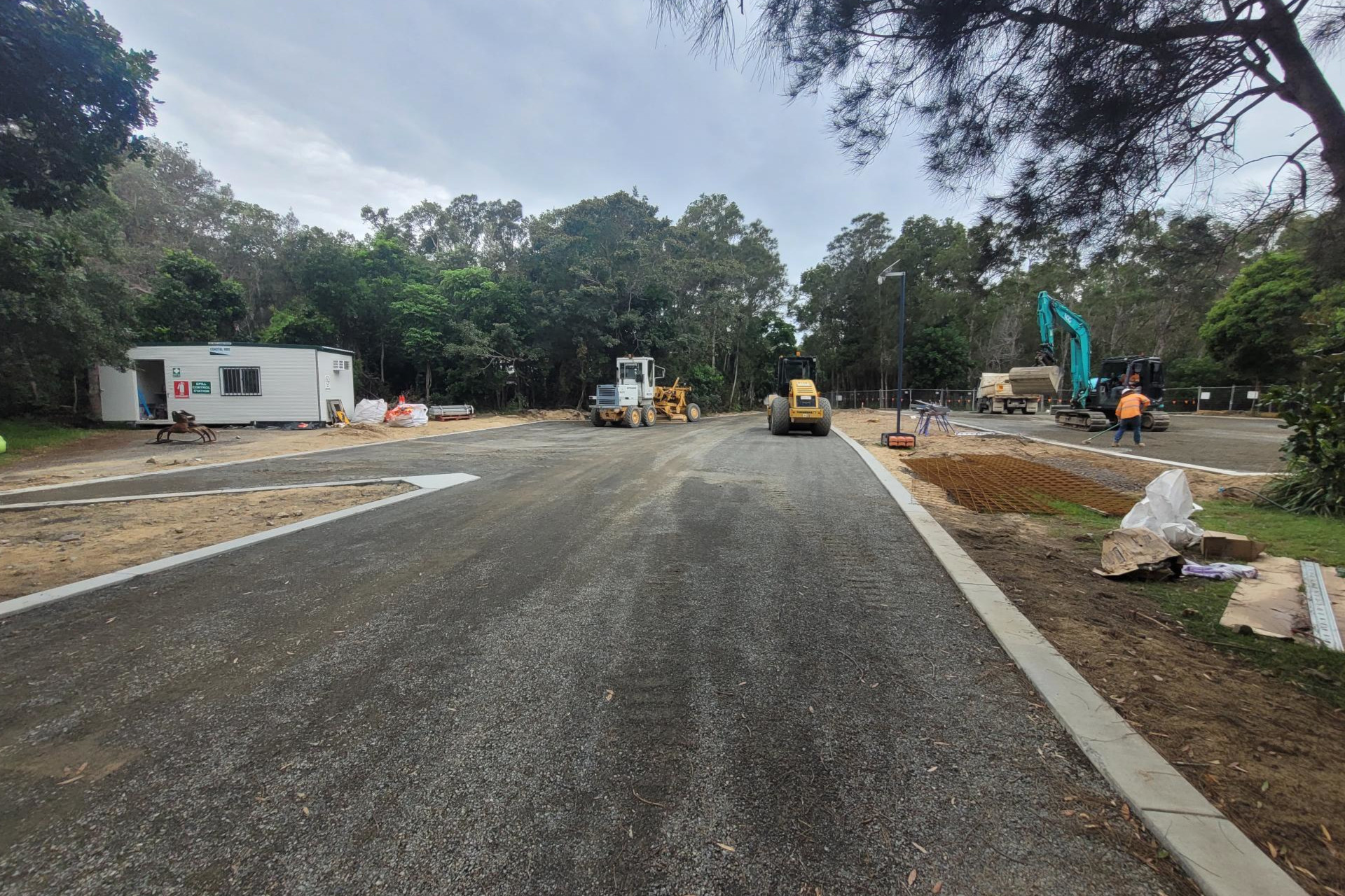 A worksite with newly laid asphalt and cement gutters. Construction tools and materials are scattered around the carpark boundaries. A site office sits on the left side.