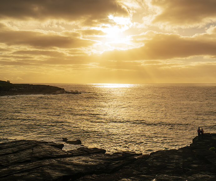 Sun setting over the ocean - two people are standing on a ledge watching whales pass by.