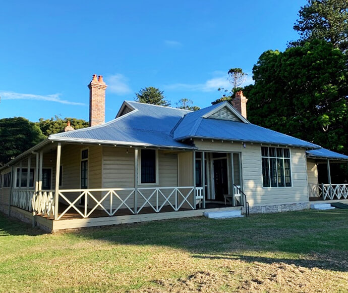 "A single-story house named 'Alpha house' with a light beige exterior and a blue metal roof. The house features a wrap-around porch with white railings and two brick chimneys. There are large windows on the front and side of the house. The house is surrounded by a grassy lawn and trees in the background. The sky is clear and blue.