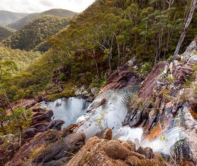 A scenic view of a natural landscape featuring a waterfall cascading down rocky terrain into a small pool of water. The surrounding area is densely forested with a variety of trees and vegetation. In the background, there are rolling hills covered in lush greenery, extending into the distance.