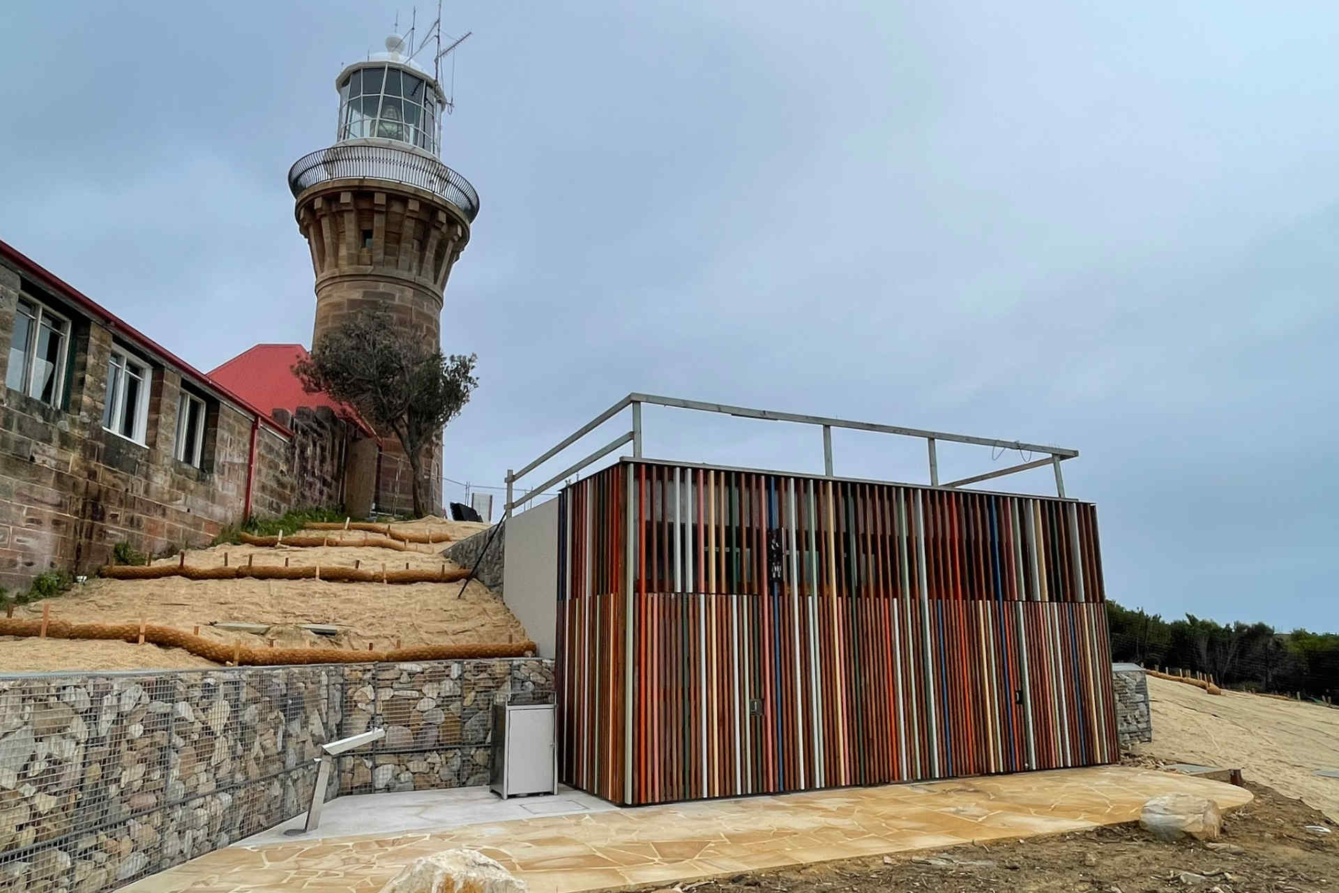 A small building with an exterior of colourful timber battens at the foot of a lighthouse under a grey sky 