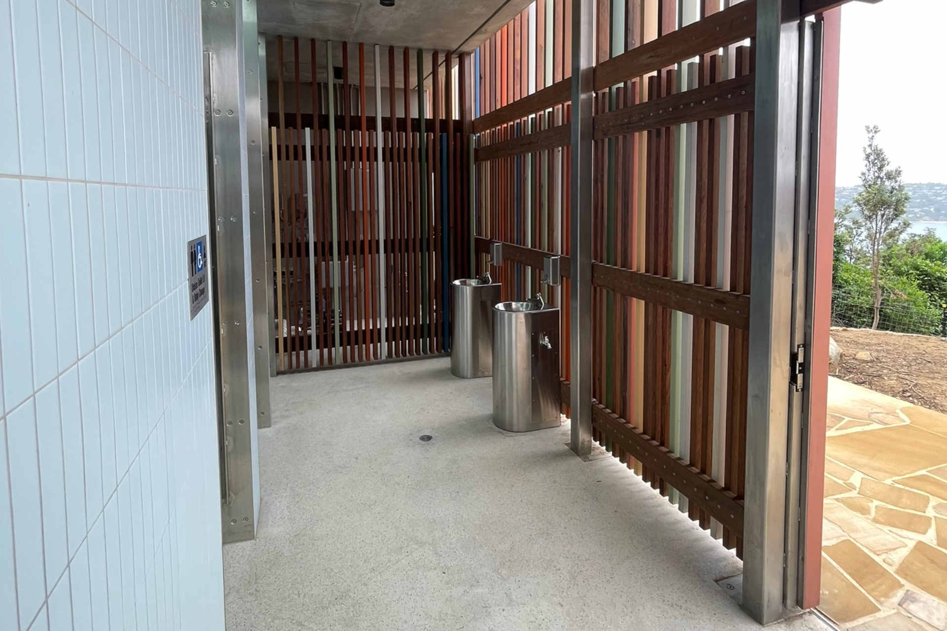 The entrance of a small amenities building with 2 small, free-standing metal wash stands and a wall comprised of multicoloured timber battens, with a glimpse of a beautiful seaside nature view through the open doorway