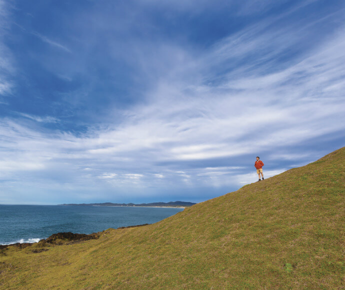 "A scenic landscape with a person standing on a grassy hill, which slopes down towards the left side of the image, leading to a coastline with the ocean visible in the background. The sky is mostly clear with some scattered clouds. The person is wearing a red jacket and white shorts, adding a sense of scale to the vast and tranquil landscape.