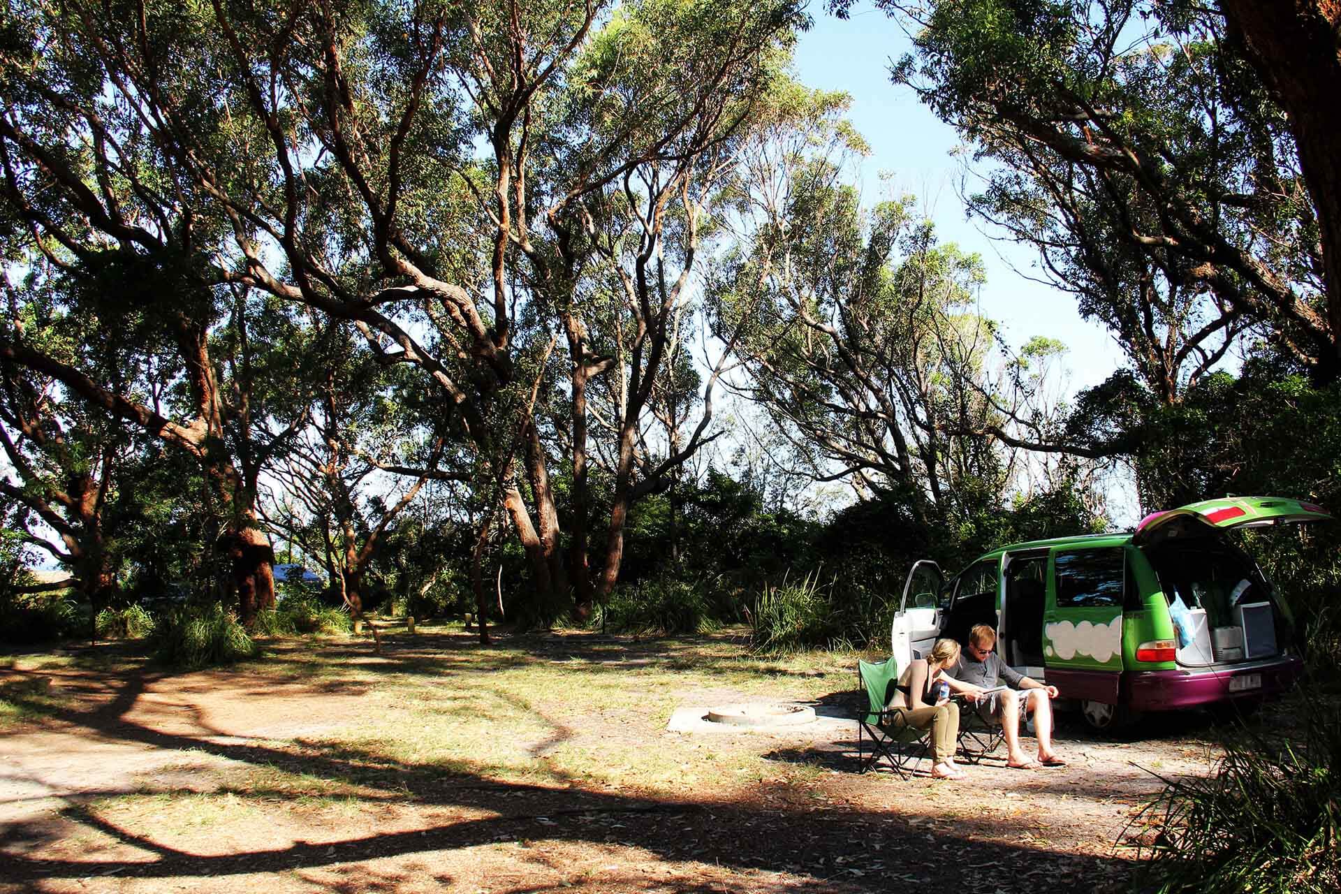 Two people in camping chairs, sitting beside a campervan, surrounded by trees