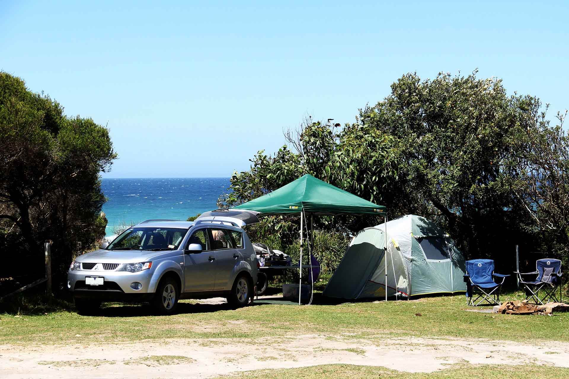 A tent, an umbrella and 2 camp chairs set up beside vehicle, water in background