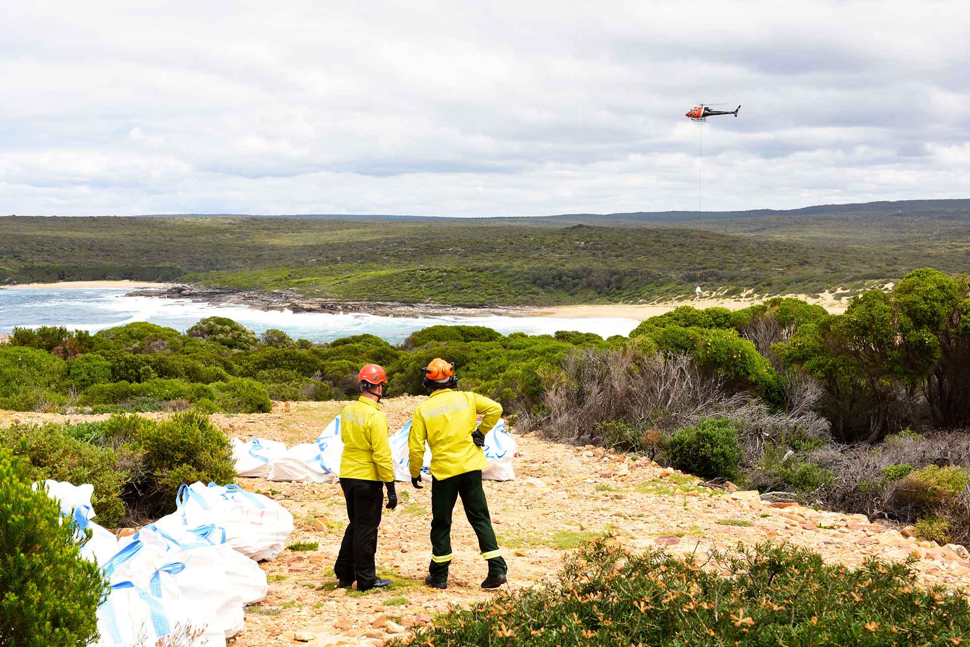 Two people in uniform standing on a rocky path with a stack of bags ncloseby, a helicopter hovers in distance