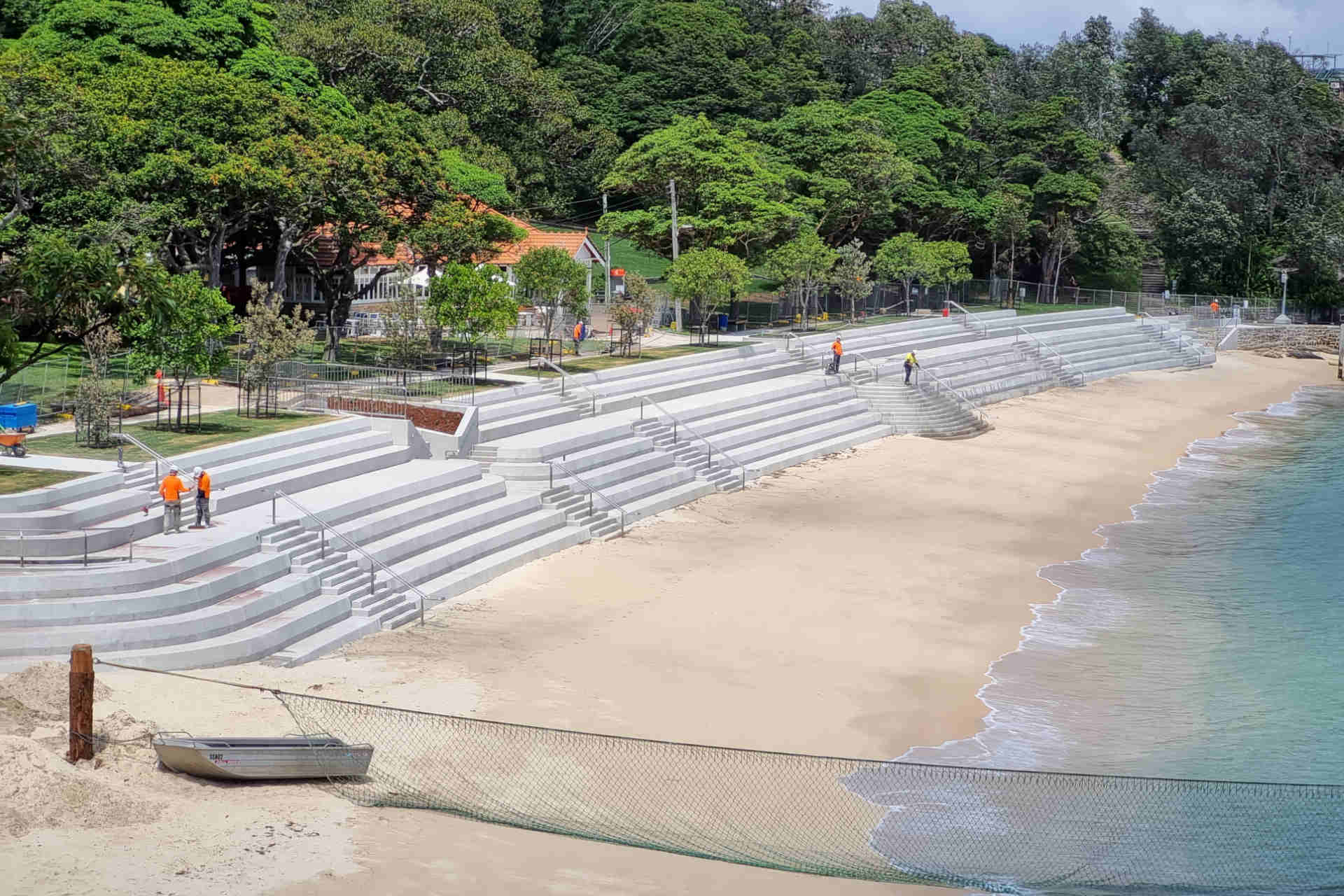 A high view of a small picturesque bay with a freshly paved wall of concrete bleachers, young saplings lining a new paved promenade along the top of the bleachers and some people in hi-vis workwear about. 