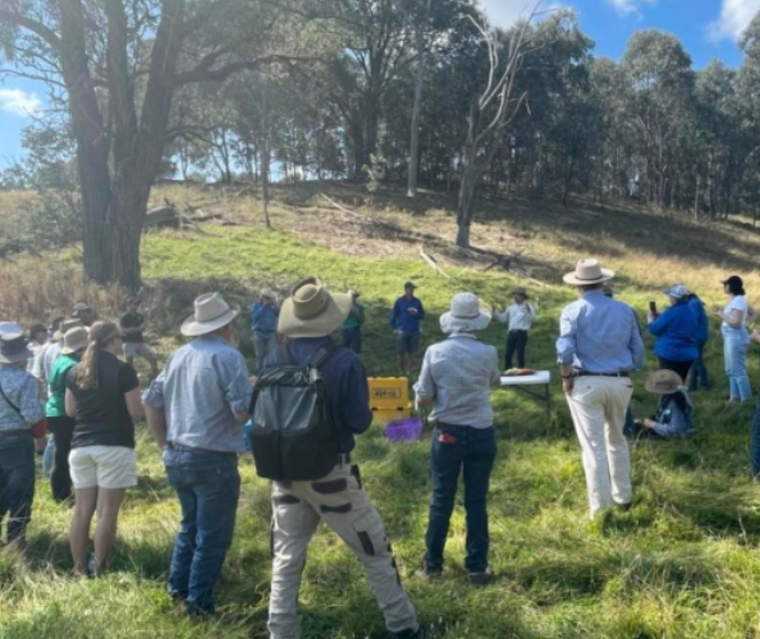 A group of people in hats gathers outdoors on a grassy area surrounded by trees, listening to a speaker. The atmosphere is relaxed and attentive.