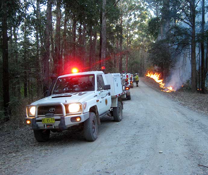 A row of utes with sirens and fire decals parked alongside a dirt road while a small grass fire burns