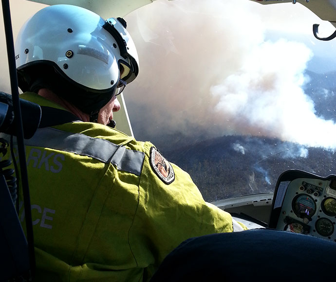 A firefighter in full uniform flying a helicopter while smoke rises up from the hazard reduction burn below