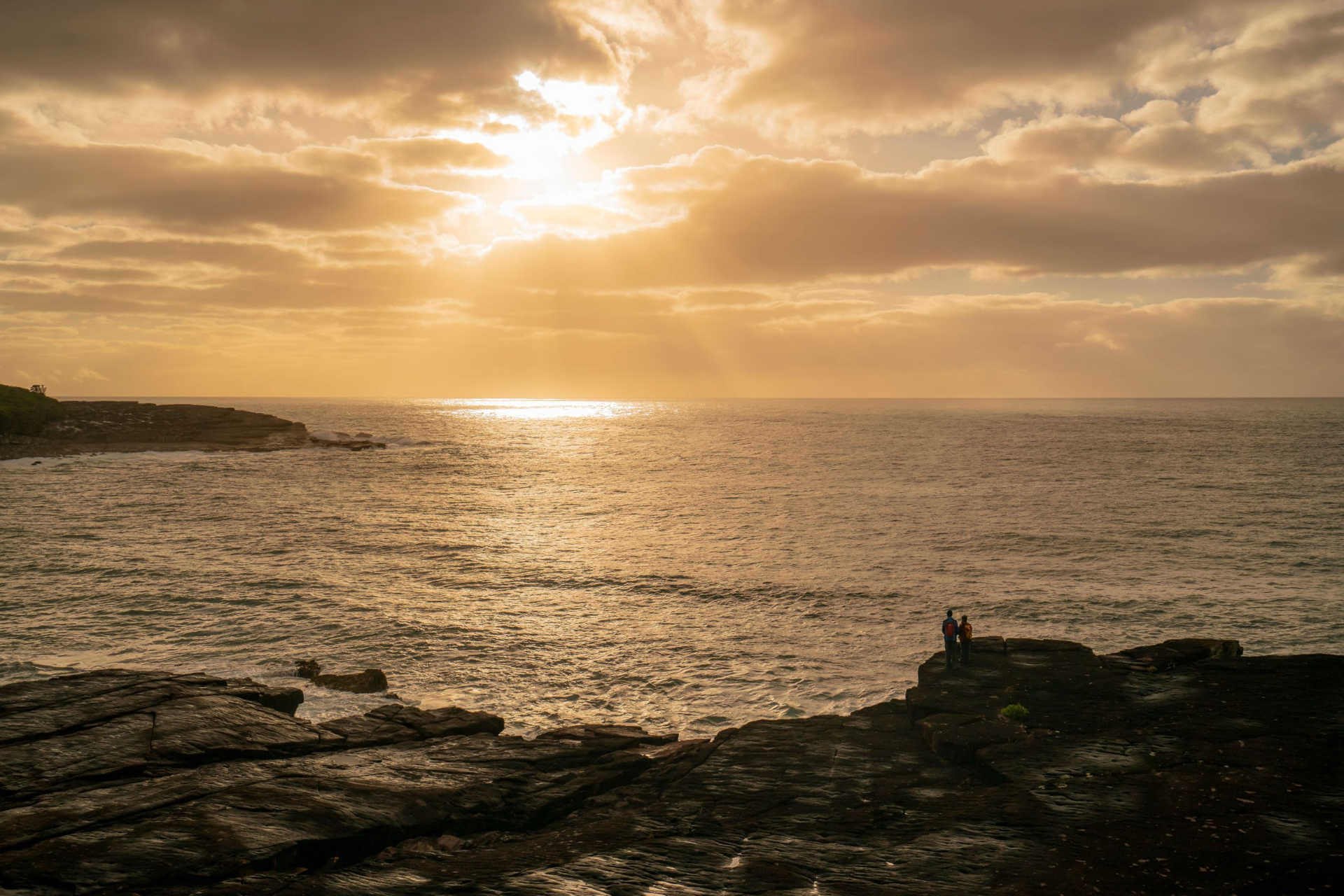 Sun setting over the ocean – two people are standing on a ledge watching whales pass by.