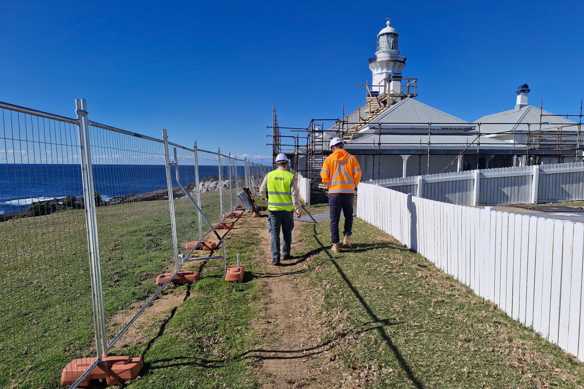 Two men in high vis clothing and helmets walk towards a cottage surrounded by scaffolding. The sea and the top of a lighthouse are visible in the distance.